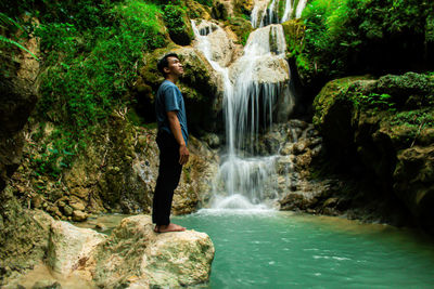 Scenic view of waterfall in rainforest with young asian man standing in profile view 