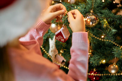 Close-up of a girl in a santa hat hangs a christmas tree toy a house on a christmas tree. 