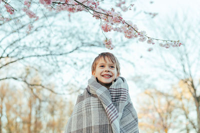 Portrait of a little boy wrapped in a blanket enjoying cherry blossoms in a city park.