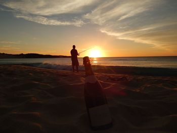 Champagne bottle by man fishing at beach against sky during sunset