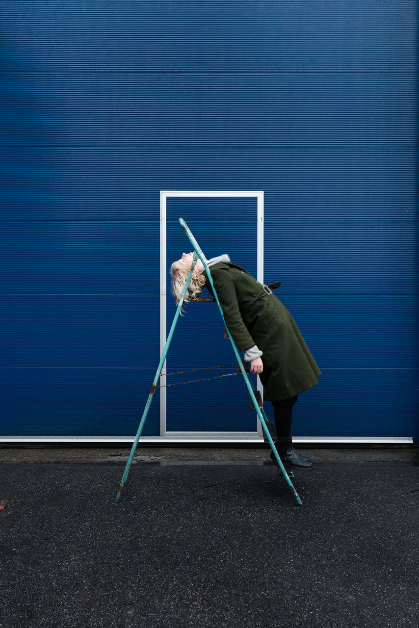 Side view full length of woman leaning on ladder by blue wall