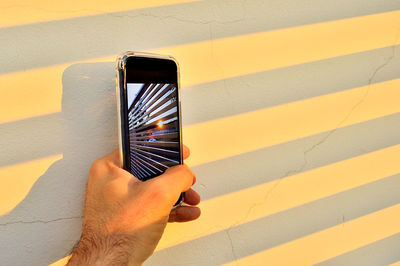 Cropped hand holding mobile phone by wall during sunset