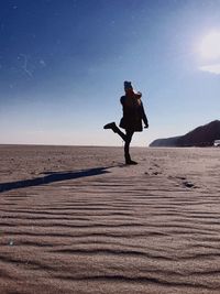 Woman standing on one leg at sandy beach against sky during sunny day