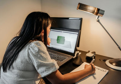 Rear view of young businesswoman sitting at desk, using laptop and 3d design software