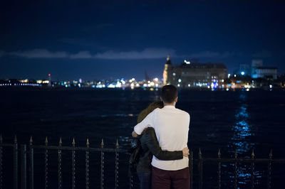 Rear view of man embracing woman while standing against sea at night