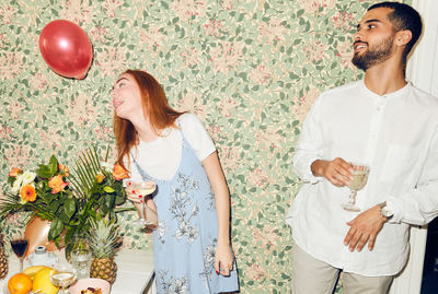 Young man looking at woman playing with balloon while standing against wallpaper at home during dinner party