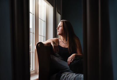 Mirror reflection of pretty woman looking out a window in a dark room.