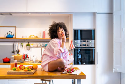 Woman eating strawberry sitting on table in kitchen at home