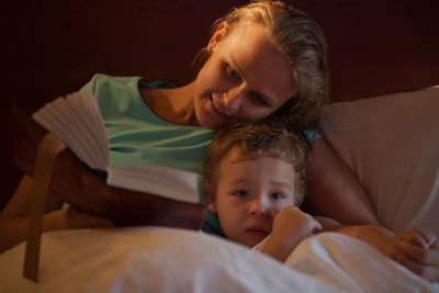 Mother holding book with son on bed in bedroom at home