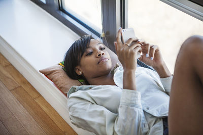 Woman using mobile phone while lying by window at home