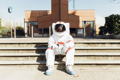 Male astronaut in space suit sitting on steps during sunny day