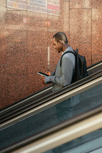 Side view of african american male worker listening to music in tws earbuds and browsing cellphone on escalator