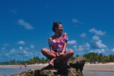 Full length of woman sitting on rock at beach against sky