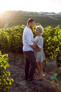 Man and a woman in white clothes stand in a grape field at sunset