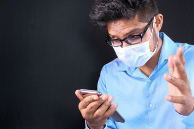 Man wearing mask gesturing while using smart phone against black background