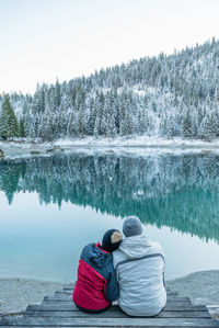 Rear view of couple sitting on lake against trees