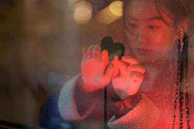 Chinese girl think of loving boyfriend drawing heart on cold fogged window covered with rain drops.