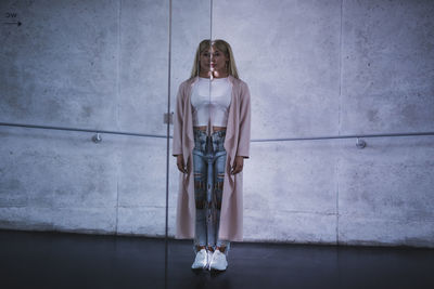 Portrait of young woman standing by mirror with reflection against wall