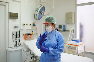 Unrecognizable female medic in respiratory mask putting on blue gloves near medical equipment in clinic