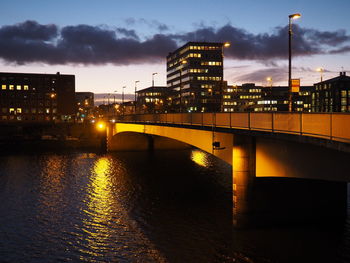 Illuminated bridge over river by buildings against sky at sunset