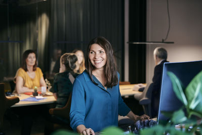 Smiling businesswoman looking away while sitting at desk against colleagues planning strategy in office