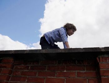 Side view of boy looking at stone wall against sky