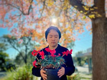 Young asian man holding red flowering cyclamen potted plant against autumn foliage, trees and sky