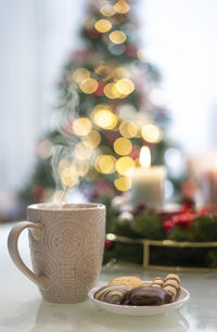 A cup of hot drink and in the background blurred candles and a christmas tree.