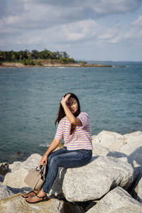 Young woman sitting on rock by sea against sky