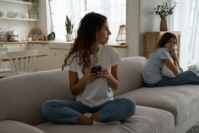 Caring puzzled mother sits on couch with phone and looks back at offended daughter after punishment