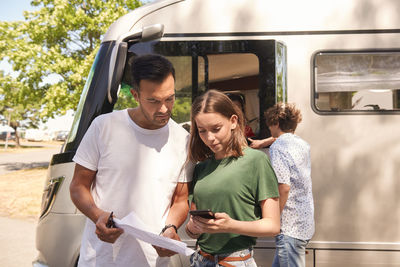 Daughter with smart phone assisting father in reading map while standing against camper van