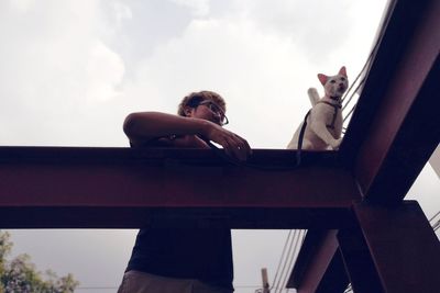 Low angle view of young man with cat on metallic railing against sky