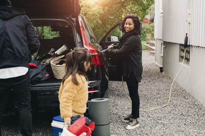 Smiling woman charging car while family loading luggage in trunk