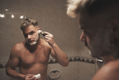 Portrait of shirtless man with arms raised in bathroom