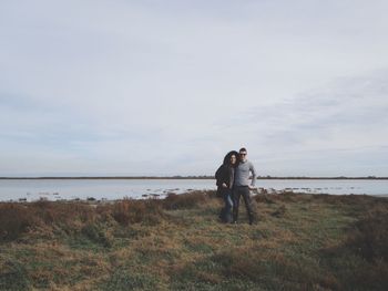 Couple standing on field by lake against sky