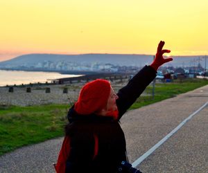 Side view of woman with hand raised standing on road against clear sky during sunset