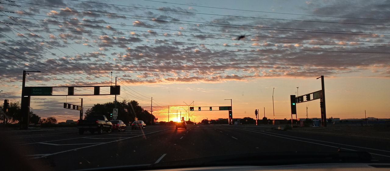sky, sunset, transportation, mode of transportation, car, evening, motor vehicle, road, dusk, cloud, land vehicle, nature, horizon, city, street, architecture, sign, orange color, afterglow, travel, windshield, sunlight, built structure, no people, light, glass, outdoors, vehicle interior, traffic, silhouette, infrastructure, car interior, on the move, highway