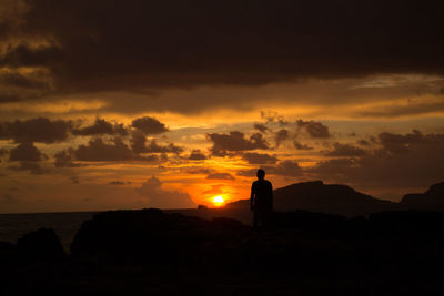 Silhouette person standing on rock against cloudy sky at sunset