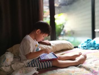 Side view of boy using digital tablet while sitting on bed by window at home