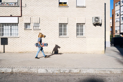 Man in horse mask walking with skateboard on footpath by building in city during sunny day