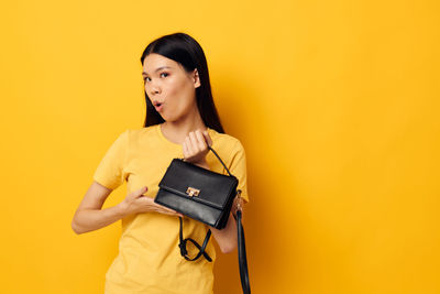 Young woman holding purse while standing against yellow background