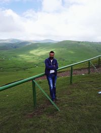 Full length portrait of man leaning on railing against grassy mountains