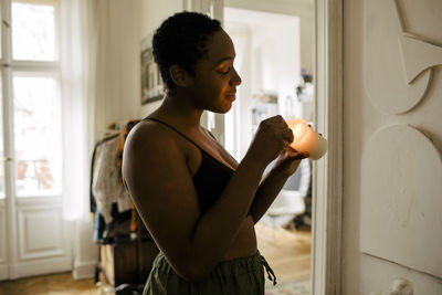 Side view of smiling young woman holding lit candle at home