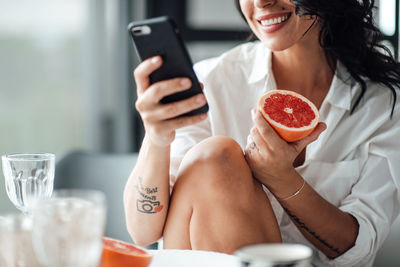 Midsection of woman holding fruit using mobile phone while sitting at home
