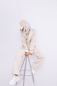 Full length of woman wearing cap sitting on stool against white background