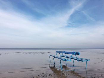Empty bench on shore against cloudy sky