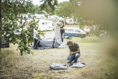 Father searching in bag while family pitching tent in background at campsite