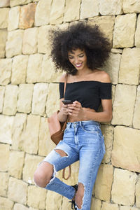 Smiling woman using mobile phone while standing against stone wall
