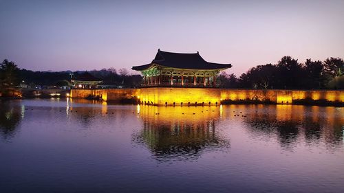 Illuminated building by lake against sky at dusk