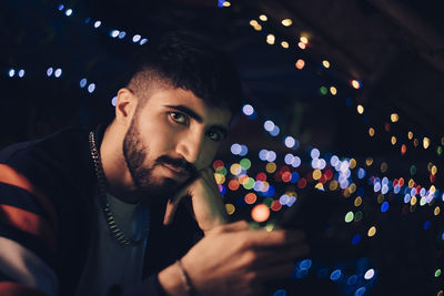 Portrait of young man holding smart phone against colorful lights at bar